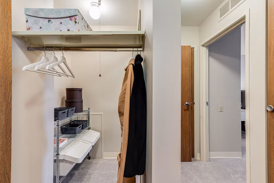 Open closet with empty hangers and coats, wire shelving holding storage bins, and a doorway leading to a carpeted room.