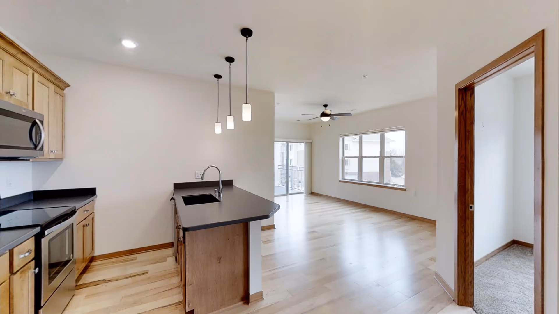 Interior view of a modern assisted living apartment featuring a kitchen with wooden cabinets, black countertops, a built-in microwave, and an island with a sink and three pendant lights. The space opens into a living area with large windows, a ceiling fan, and light wood flooring. A doorway on the right leads to a carpeted room.