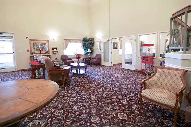A spacious common area with patterned carpet, several upholstered chairs and small tables, a large window with curtains, and a staircase with a wooden railing. The room has light-colored walls and is well-lit with wall sconces and natural light.