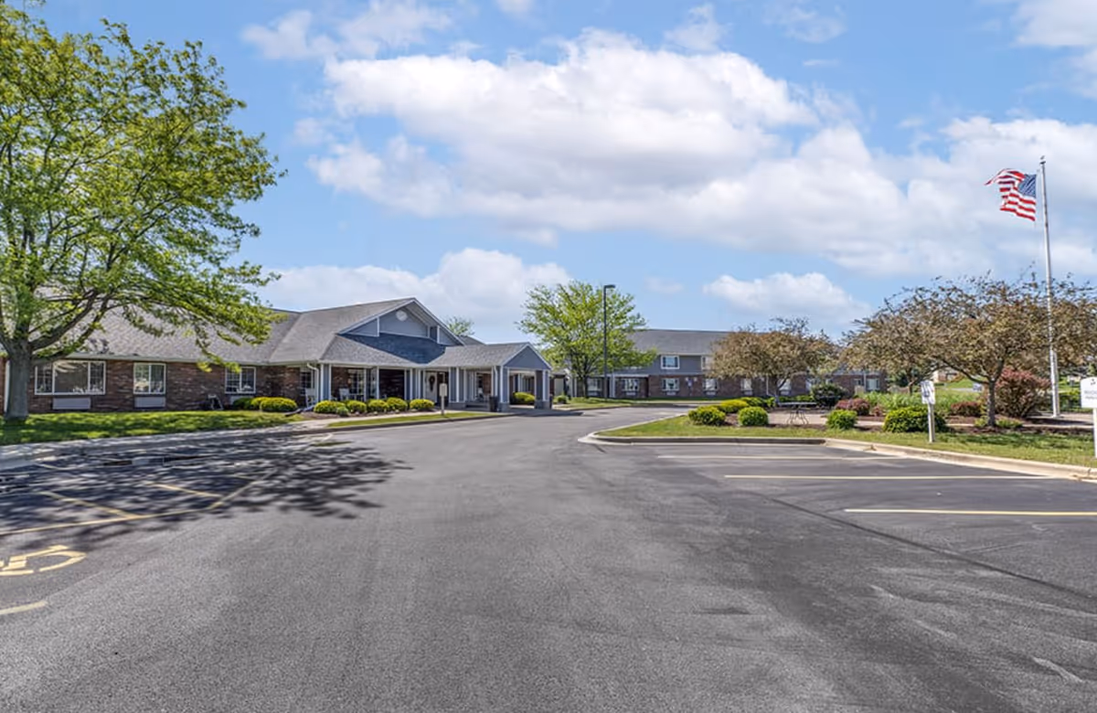 Front entrance and parking lot of a single-story senior living facility with landscaped grounds and an American flag.
