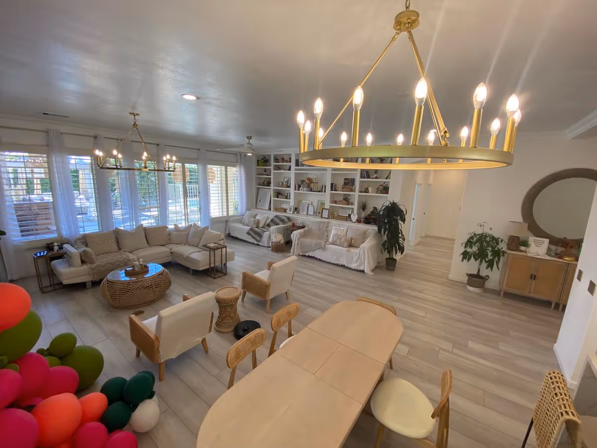 A spacious and well-lit living room area with large windows covered by sheer white curtains. The room features a large beige sectional sofa, two armchairs, a round wicker coffee table, and built-in white shelves filled with decorative items and books. There are two modern chandeliers hanging from the ceiling, a wooden dining table with chairs in the foreground, and several potted plants adding greenery to the space.