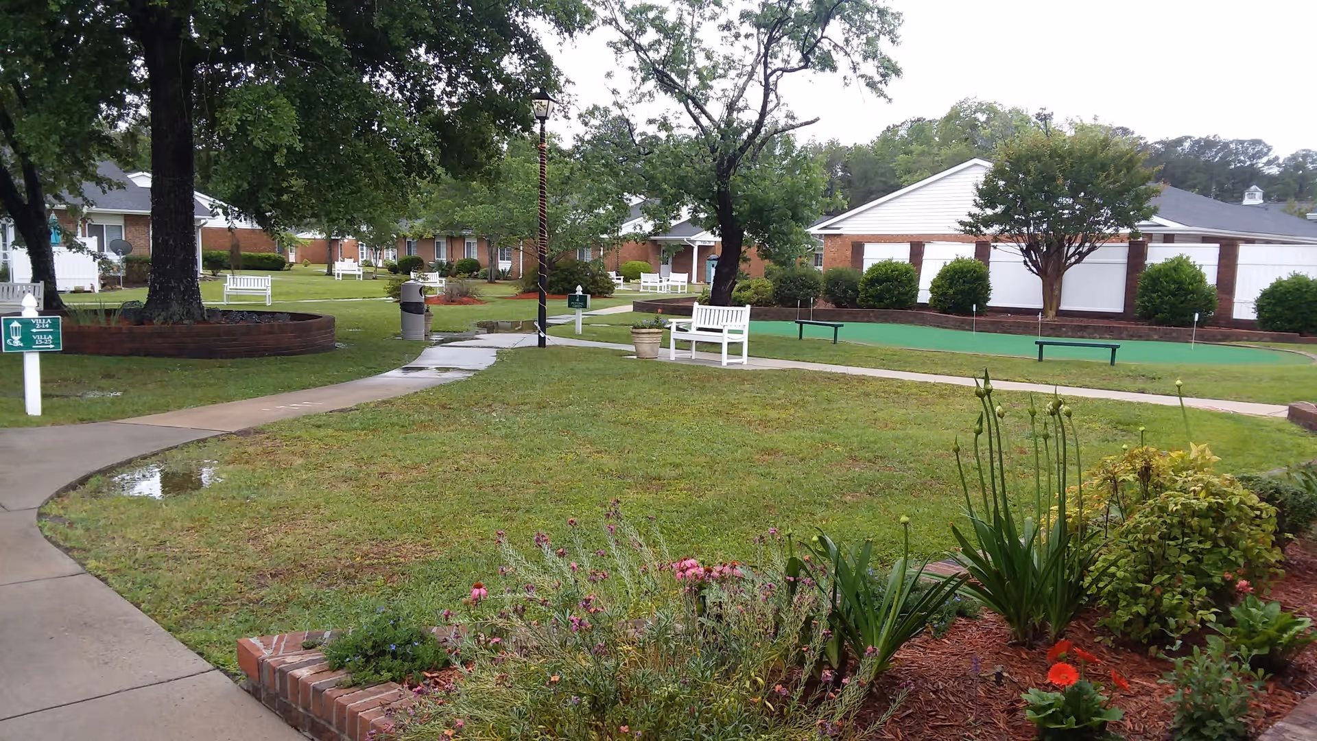 Outdoor garden area in a senior living facility with green grass, flower beds, several white benches, trees, and a paved walking path. Residential-style buildings are visible in the background under a cloudy sky.