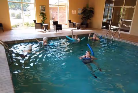 Several elderly residents using pool noodles while exercising in an indoor swimming pool with seating along the far wall.