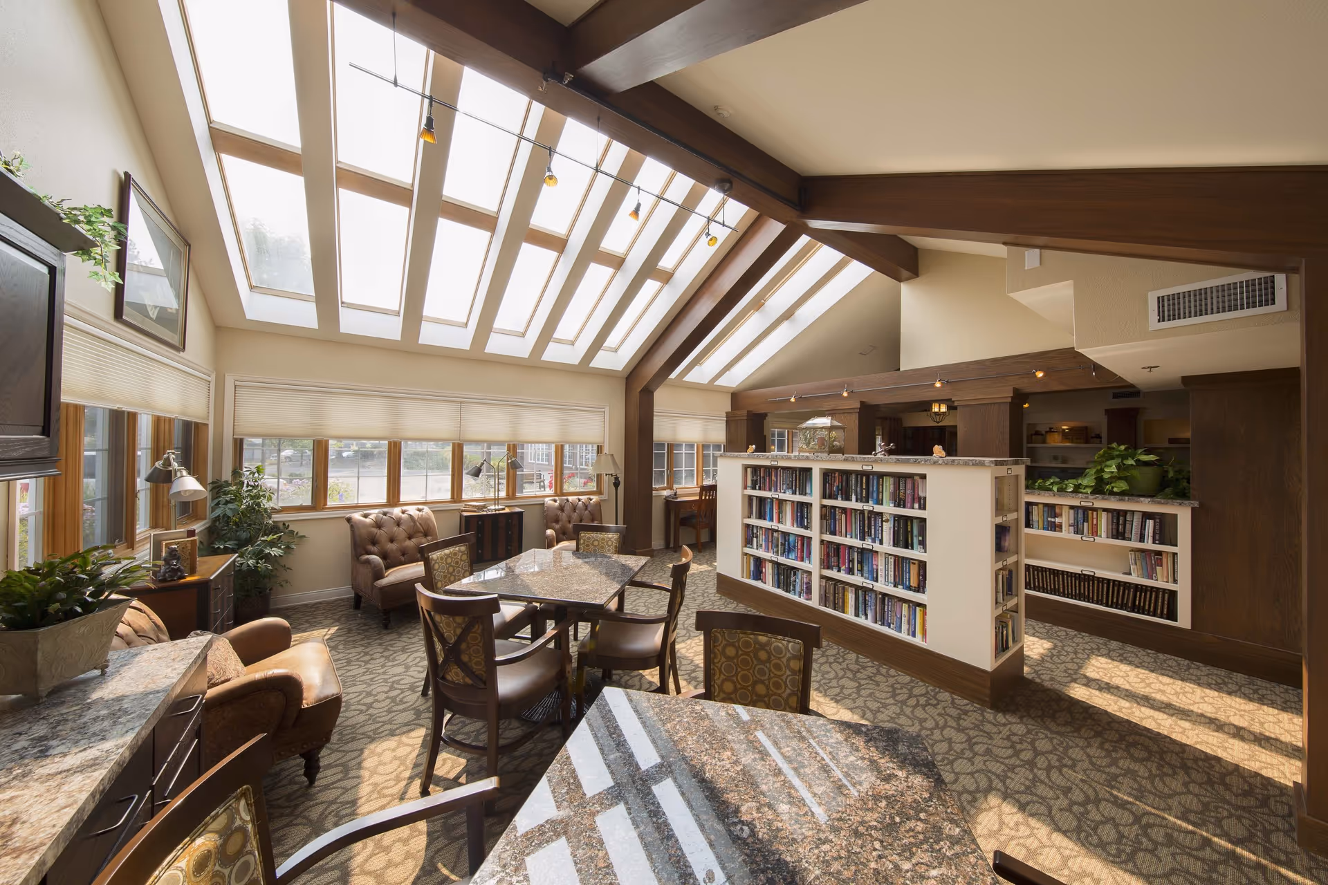 A bright and spacious common area with large skylights and windows letting in natural light. The room features comfortable leather armchairs, tables with patterned chairs, bookshelves filled with books, and several potted plants. The carpet has a decorative pattern and the ceiling has wooden beams with track lighting.