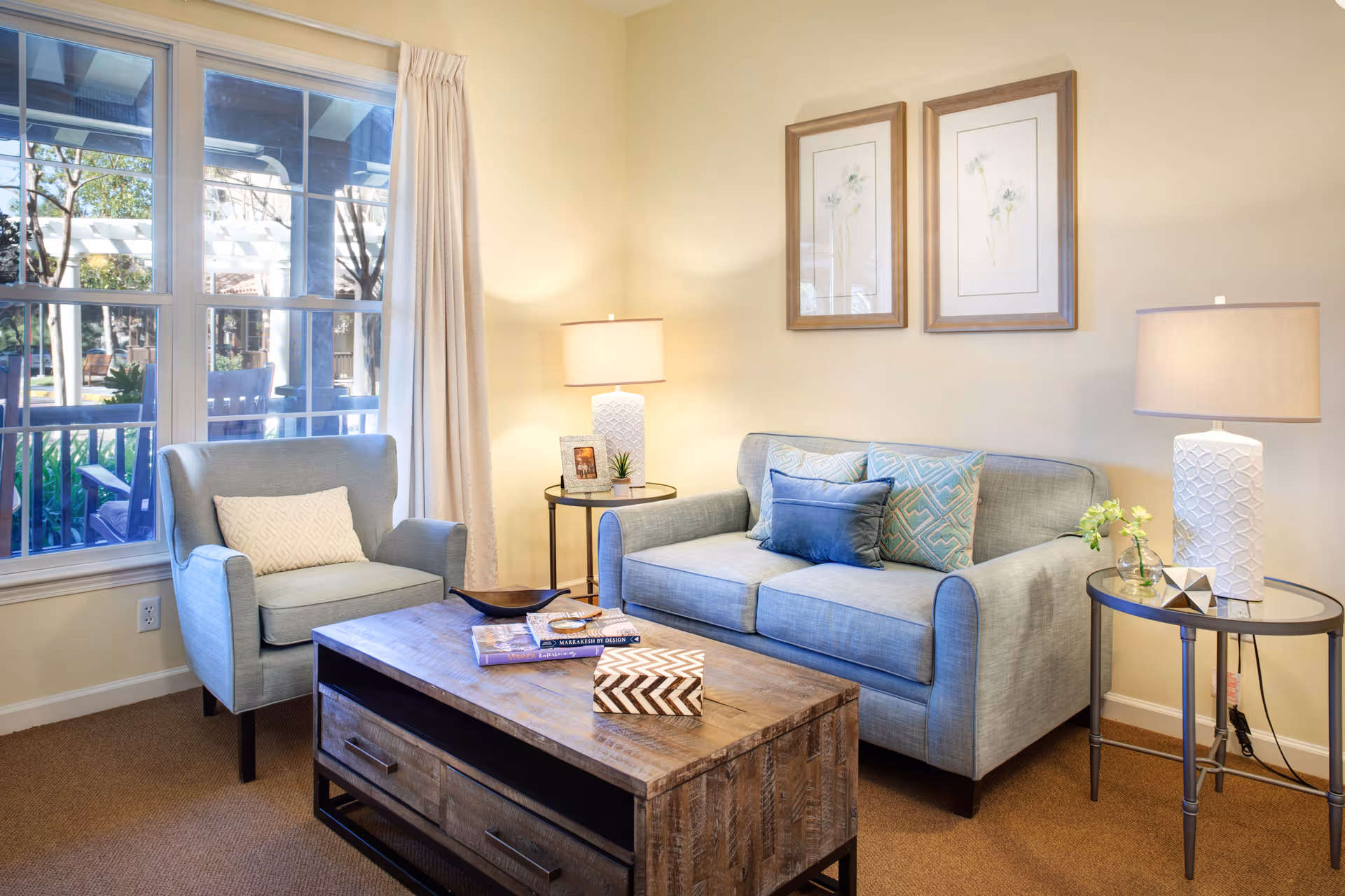 Well-lit living room with a sofa, armchair, wooden coffee table, and lamps beside a large window.