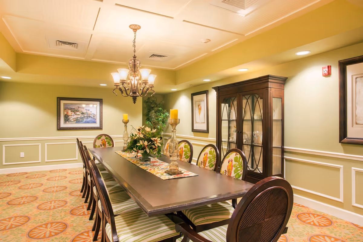 A well-lit dining room with a long dark wooden table surrounded by chairs with patterned cushions. The table is decorated with a floral centerpiece and two large candle holders. The walls are painted light green with white trim, and there are framed pictures and a glass-fronted cabinet displaying glassware. A chandelier hangs above the table, and the carpet has a colorful circular pattern.