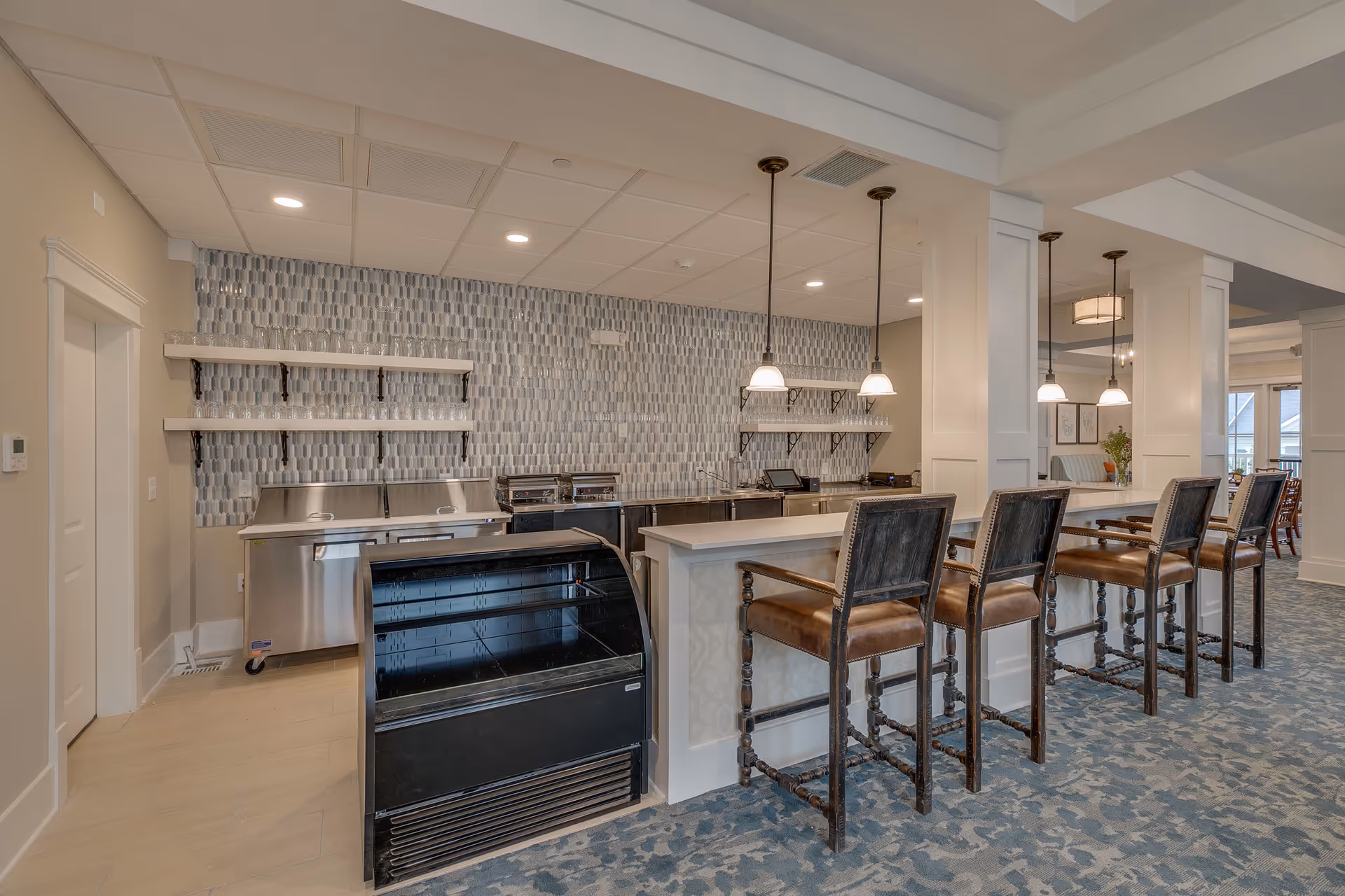 Interior view of a modern kitchen area in a senior living facility featuring a long counter with four high wooden chairs with leather seats. The back wall has a patterned tile backsplash, open shelves, and stainless steel appliances. Pendant lights hang above the counter, and the floor is carpeted with a blue patterned design.