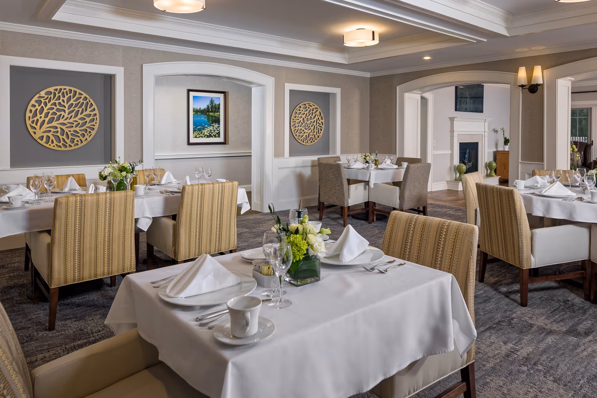 A well-lit dining room with multiple tables covered in white tablecloths, each set with white napkins, plates, glasses, and silverware. The room features beige patterned chairs, decorative wall art, a framed landscape painting, and a fireplace in the background.