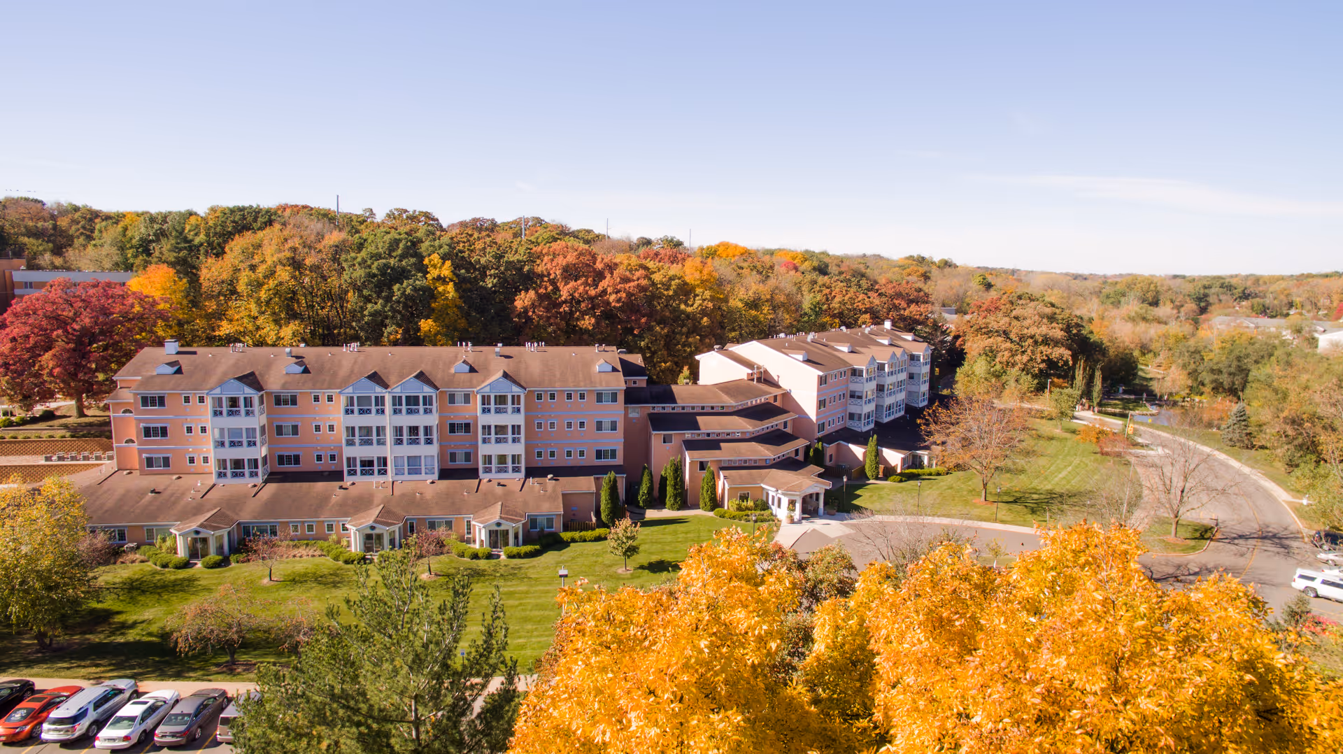 Aerial view of a multi-story senior living building set in landscaped grounds with autumn trees, lawns, a parking area and a curved driveway.