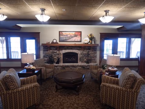 A cozy living room area with four patterned armchairs arranged around a dark brown octagonal ottoman. The room features a stone fireplace with a wooden mantle, decorated with a clock, a framed picture, and a vase with flowers. Two table lamps with beige shades sit on side tables between the chairs. Large windows with wooden frames allow natural light into the room.