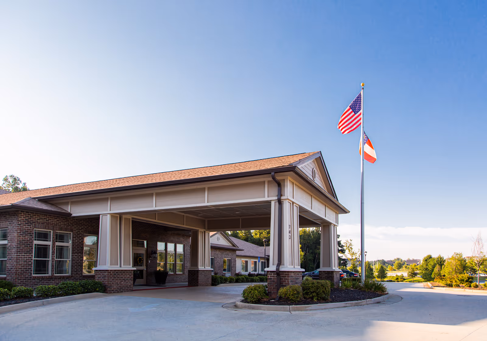 Exterior view of Marshall Pines Assisted Living & Memory Care building entrance with a covered drop-off area, brick walls, and two flagpoles displaying the American flag and another flag. The sky is clear and blue with some greenery in the background.