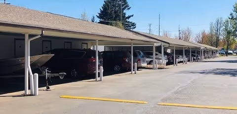 Covered parking area with multiple cars and a boat parked under a long roofed structure, with trees and power lines visible in the background.