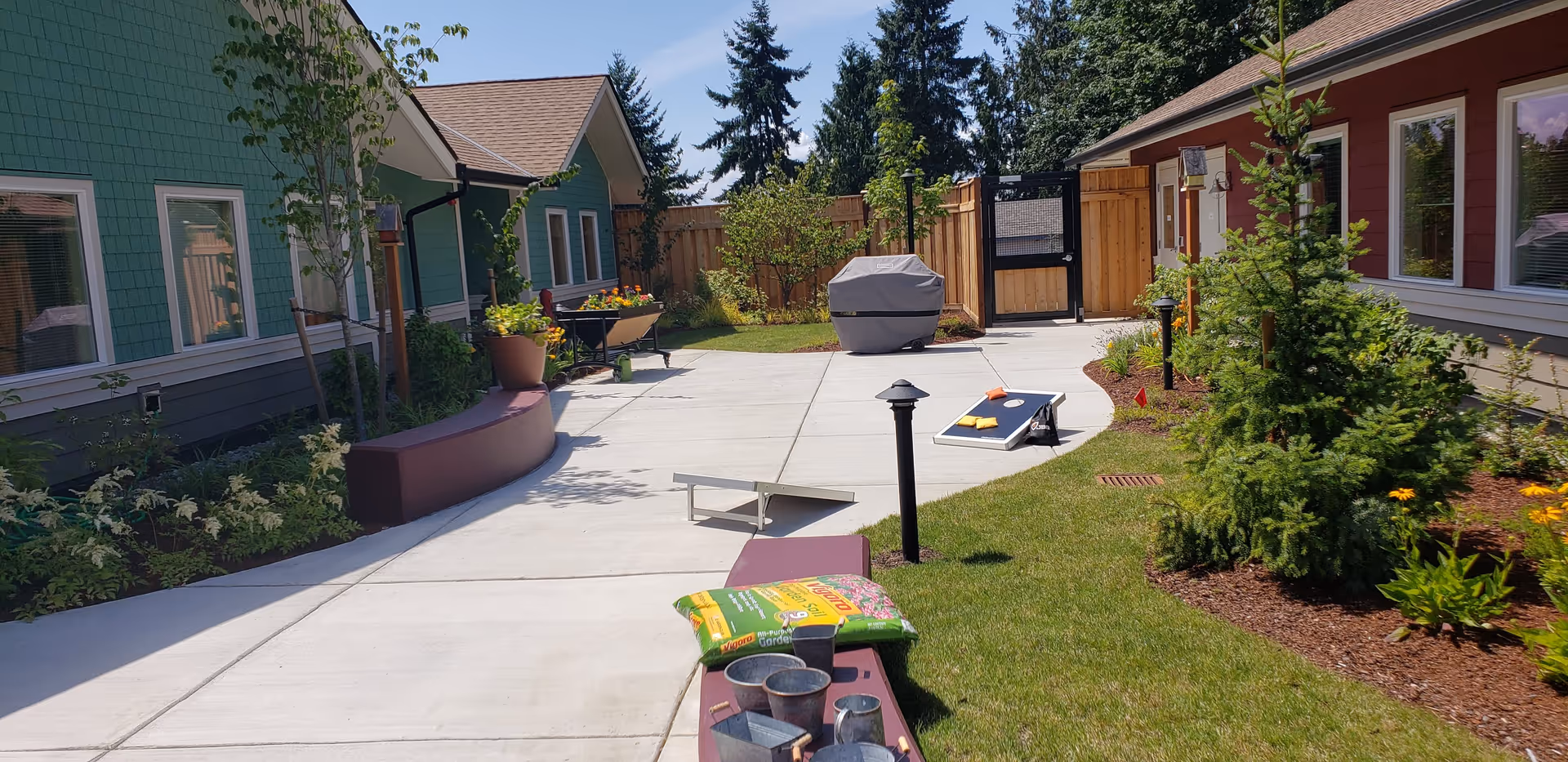 Outdoor courtyard area between two single-story buildings with green and red siding. The courtyard has a concrete walkway, garden beds with plants and flowers, a covered grill, a cornhole game set, and gardening supplies on a curved bench. Trees and a wooden fence surround the area under a clear blue sky.