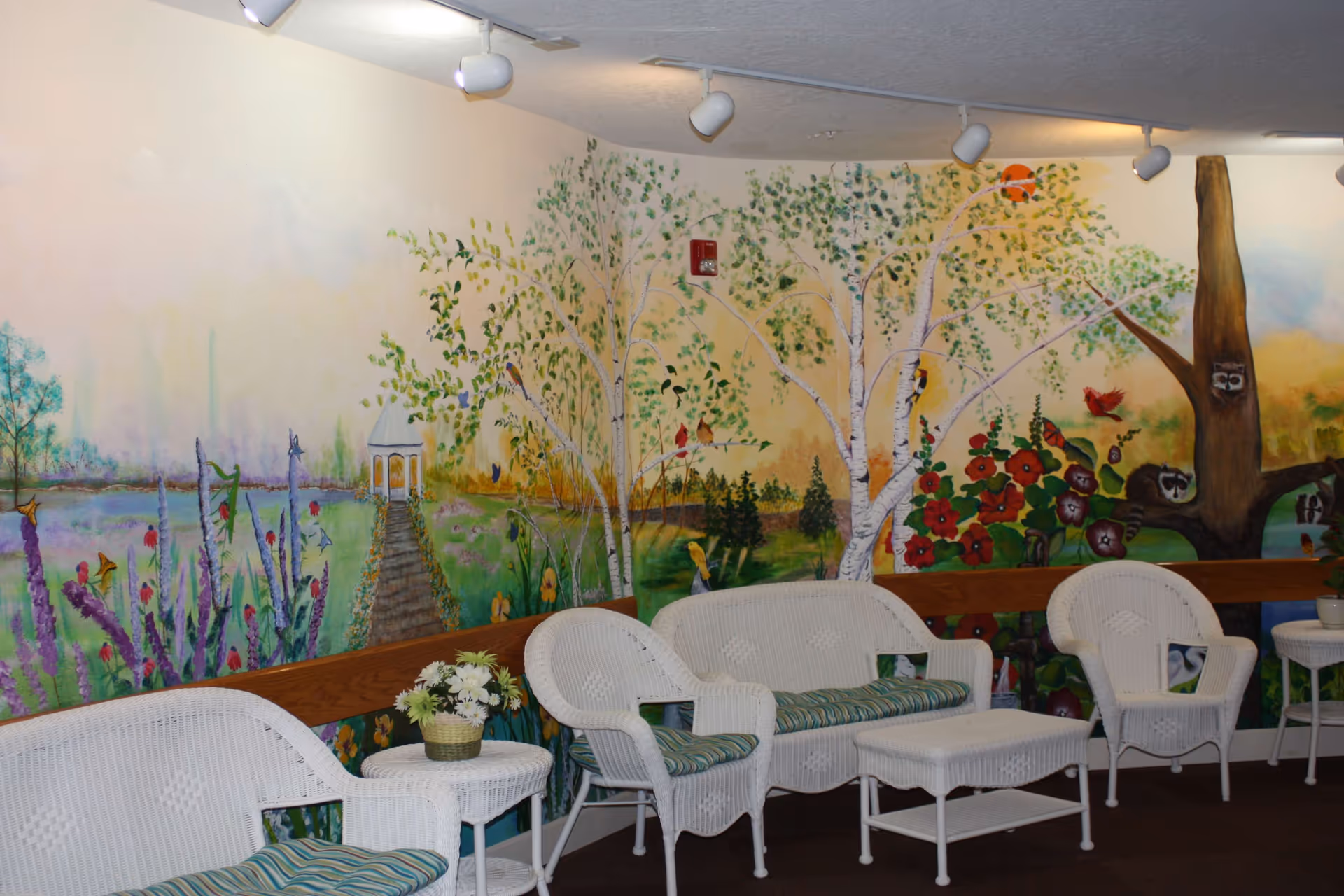A cozy indoor seating area with white wicker furniture including chairs, a loveseat, a coffee table, and side tables. The furniture has green and blue striped cushions. The walls feature a colorful mural depicting a garden scene with trees, flowers, birds, raccoons, and a gazebo in the background. A basket of white flowers is placed on one of the side tables.