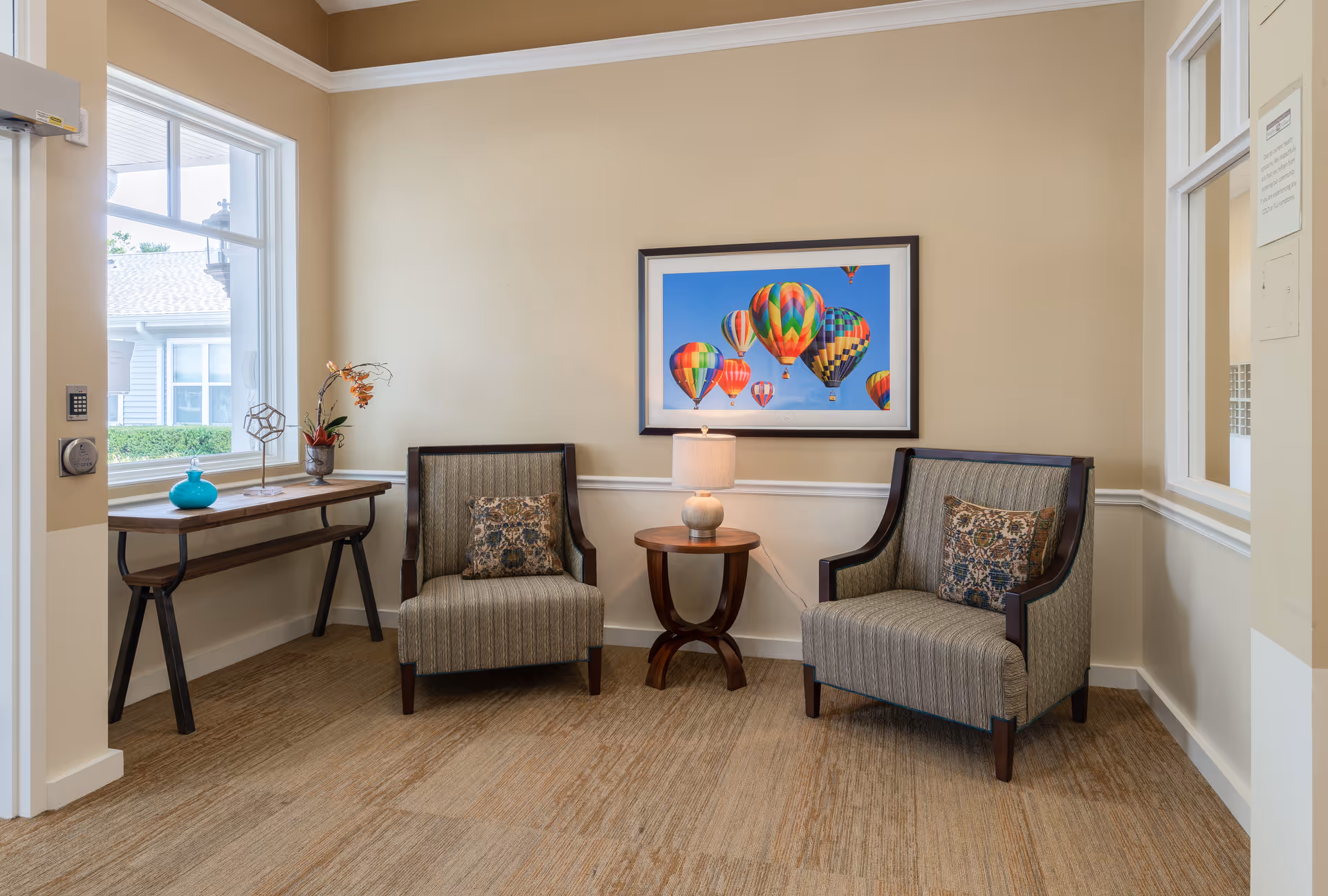 A cozy seating area in a senior living facility with two upholstered armchairs featuring patterned cushions, a small round wooden table with a lamp between them, a console table with decorative items including a blue vase and a plant, and a colorful framed picture of hot air balloons on the beige wall.