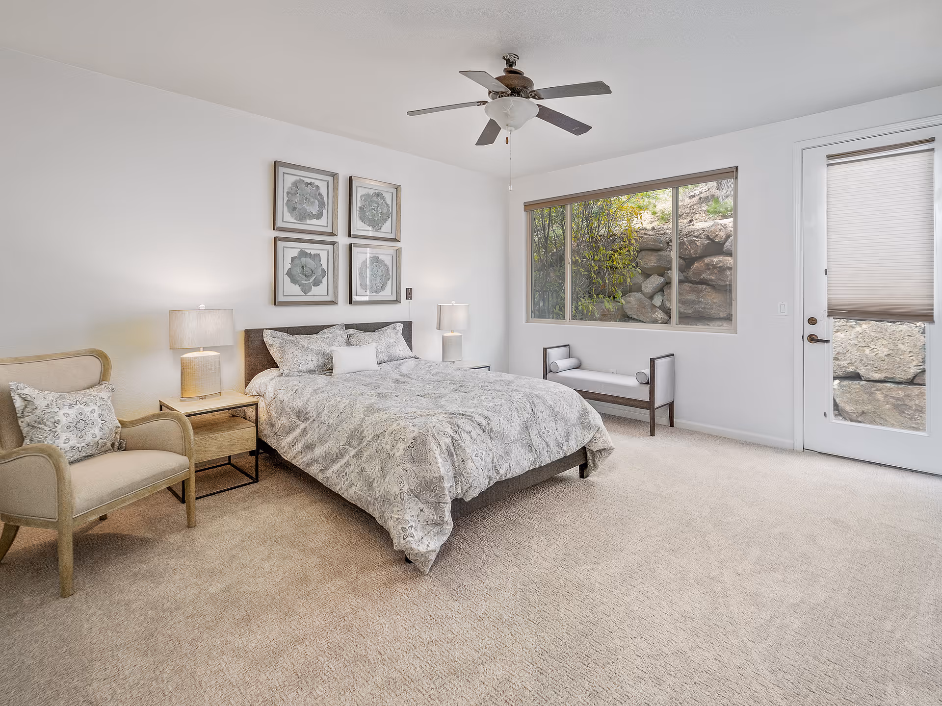 Spacious neutral bedroom with a bed flanked by nightstands and lamps, an armchair, a bench, a ceiling fan, and a large window and door to the outside.