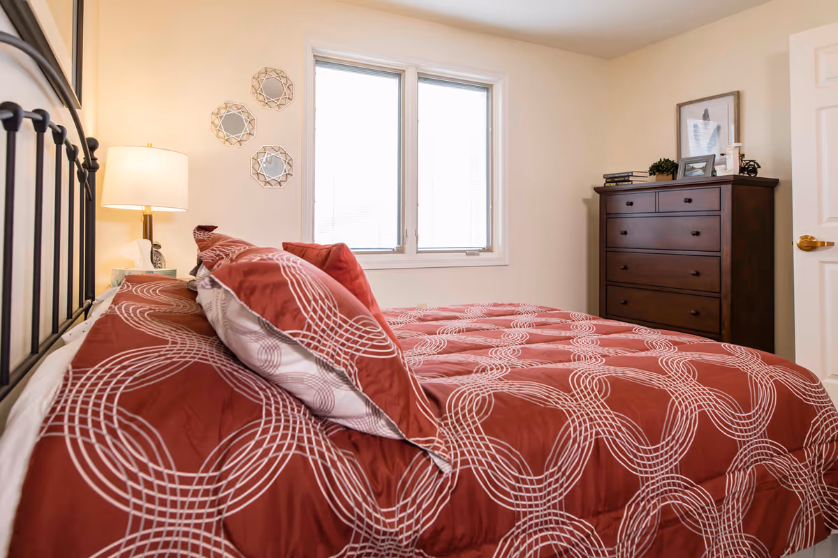A bright bedroom featuring a bed with red patterned bedding, a bedside lamp, decorative wall mirrors, and a dark wood chest of drawers beneath a window.