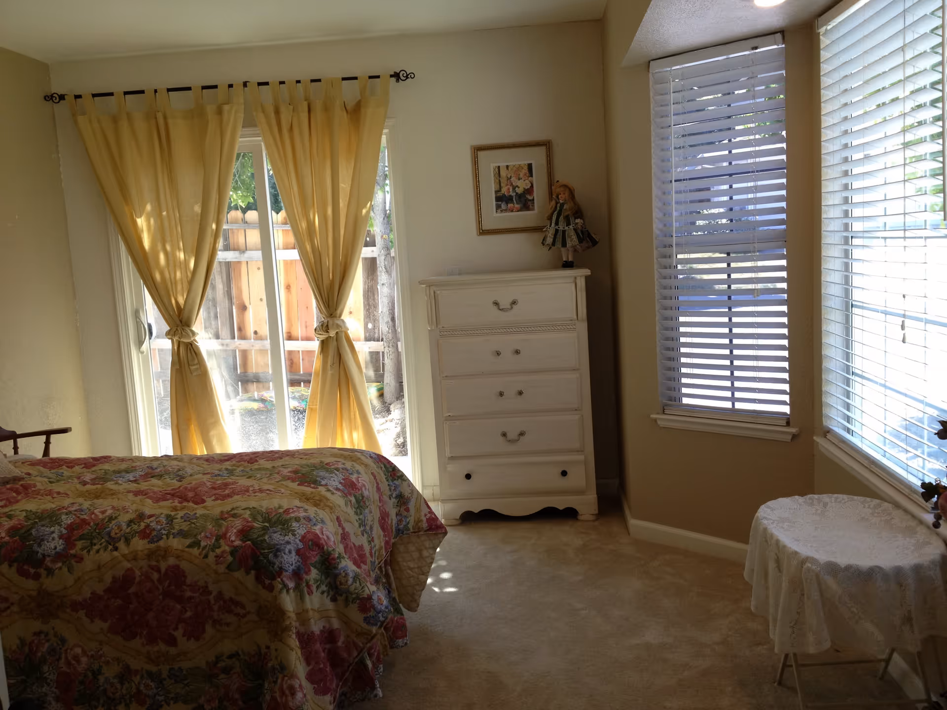 A bedroom with a bed covered in a floral patterned bedspread, a white chest of drawers with a decorative doll and framed picture on top, yellow curtains tied back over a sliding glass door, and two windows with white blinds. There is a small round table covered with a white lace tablecloth near the windows.