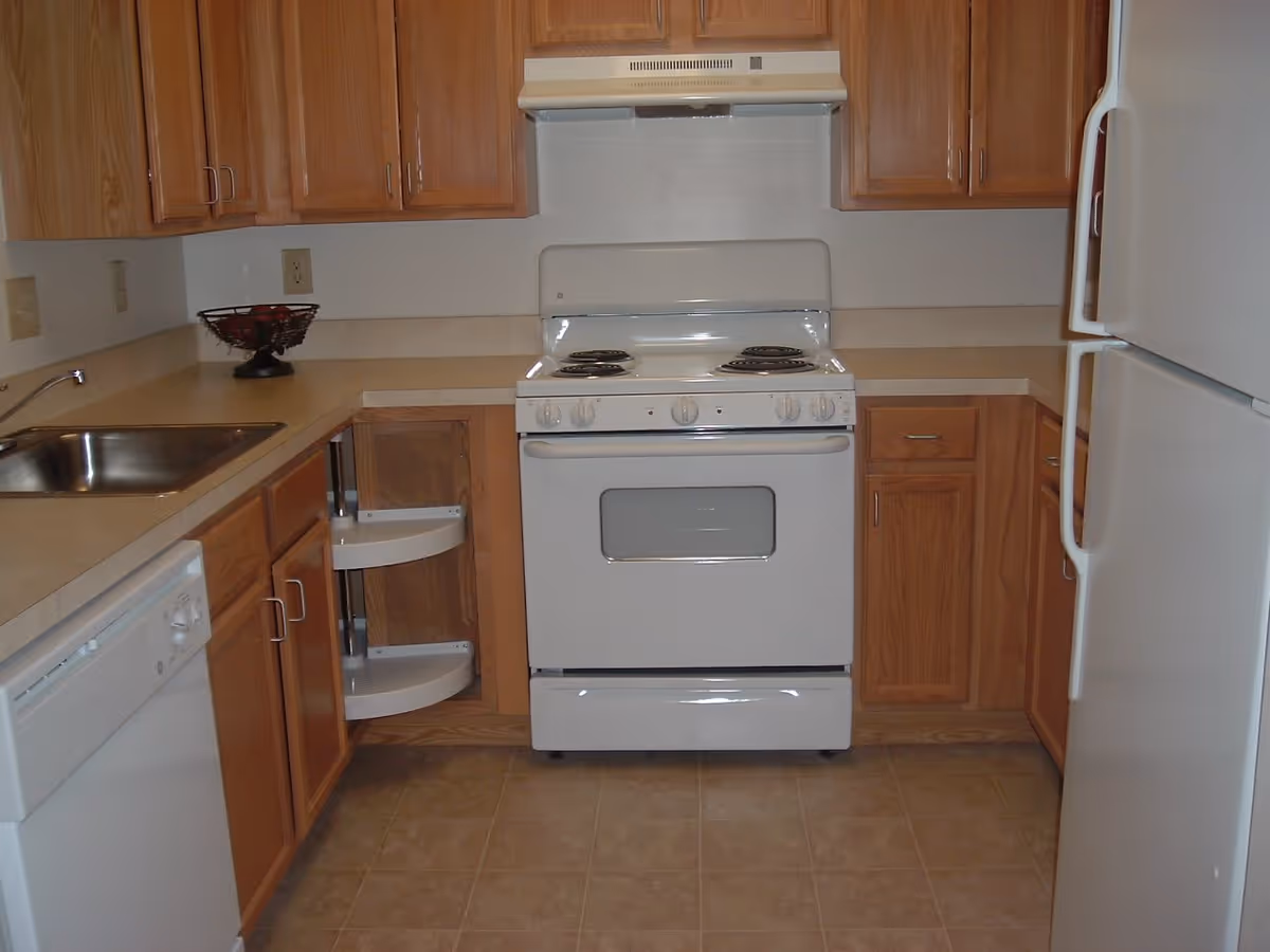 Compact kitchen with a white stove, refrigerator and dishwasher, wooden cabinets, sink, and corner pull-out shelving.