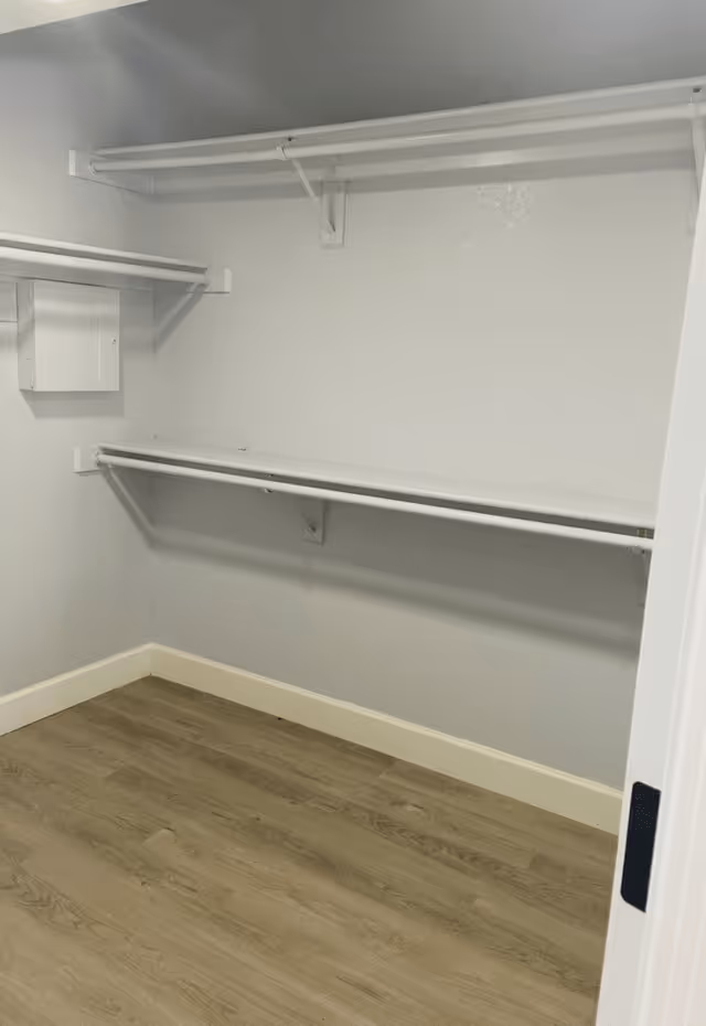 Empty walk-in closet with light gray walls, white shelving and hanging rods, and wood-look flooring.