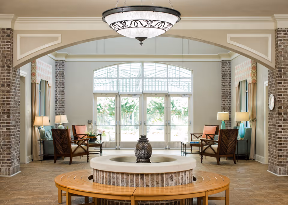 A bright and spacious common area in a senior living facility featuring a circular stone water fountain with a wooden bench surrounding it. The room has large glass doors letting in natural light, brick pillars, and comfortable seating with armchairs and side tables with lamps on either side. A decorative chandelier hangs from the ceiling.