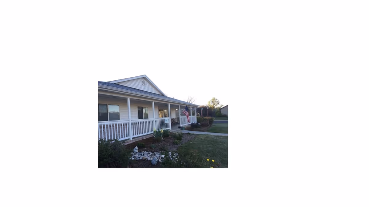Exterior view of a single-story building with a covered porch featuring white railings, an American flag, and a landscaped garden with bushes and rocks in front.