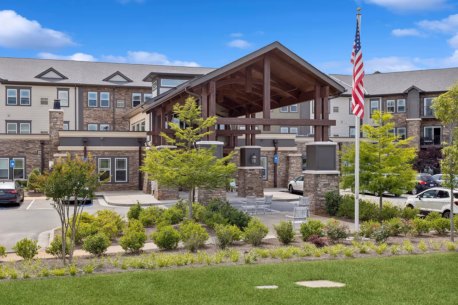 Exterior view of The Phoenix at Union Hill senior living facility showing a large covered entrance with wooden beams, surrounded by landscaped greenery, small trees, and an American flag on a flagpole. Several cars are parked nearby under a partly cloudy sky.