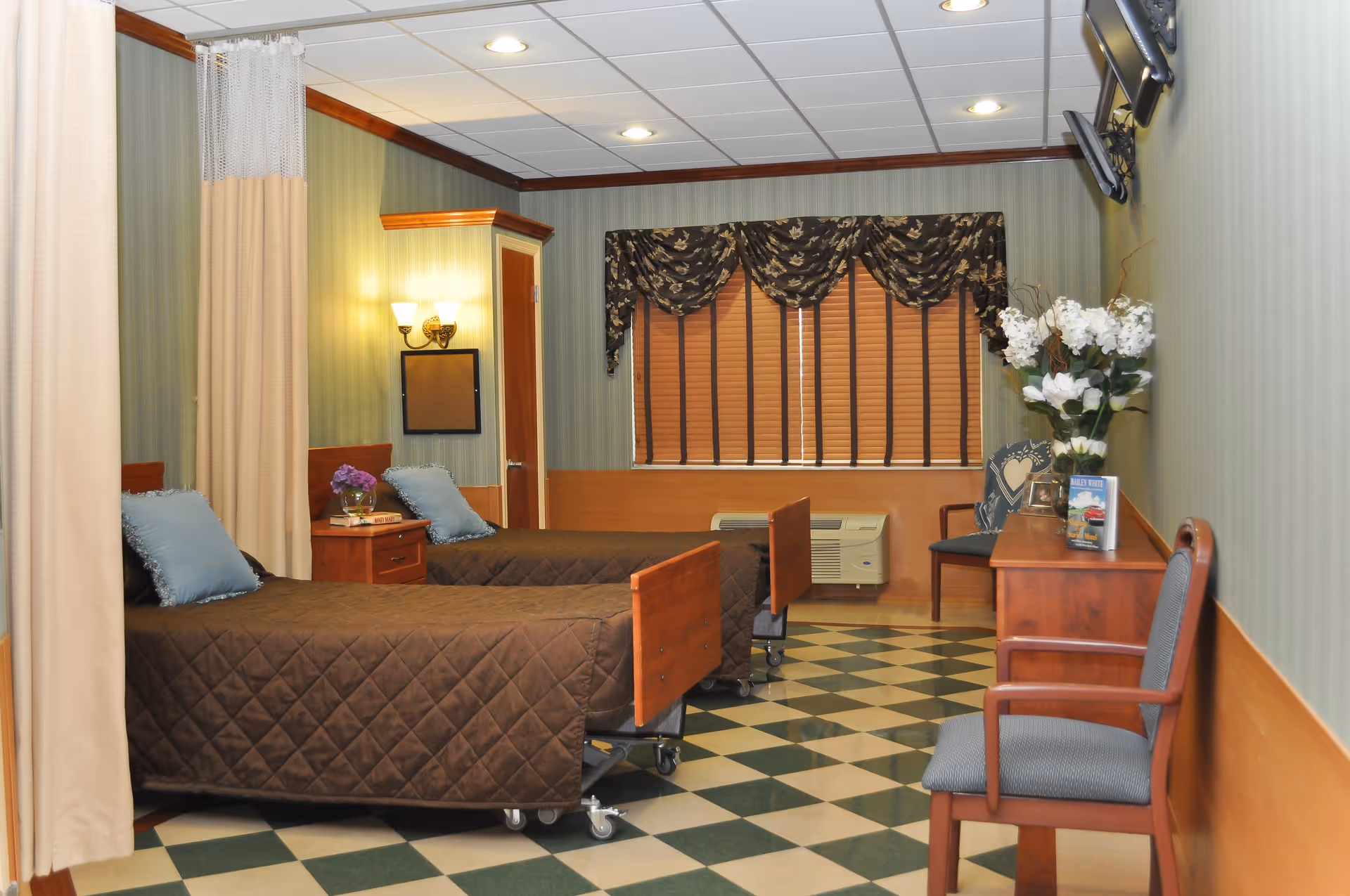 A room with two single beds covered in brown quilted bedspreads, each with a blue pillow. There is a wooden nightstand between the beds with a small flower arrangement. The room has green striped wallpaper, a checkered green and beige floor, and a window with closed blinds and a dark floral valance. A wooden dresser with a vase of white flowers and a chair are on the right side, and a TV is mounted on the wall above the dresser. A curtain partially divides the beds.