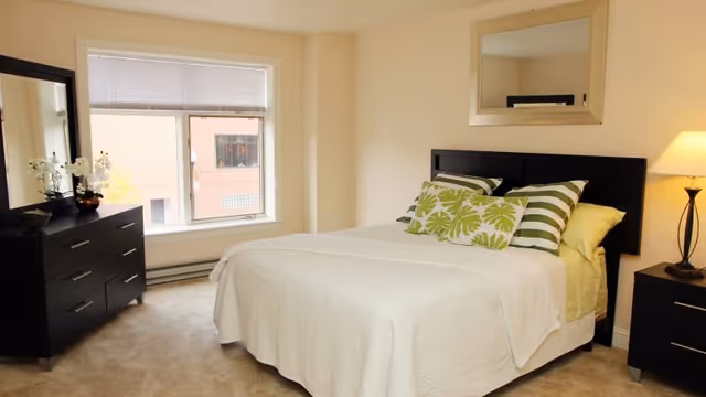 A bright bedroom with a large window, a bed with white bedding and green patterned pillows, a black dresser with a mirror and decorative flowers, a nightstand with a lamp, and a wall mirror above the bed.