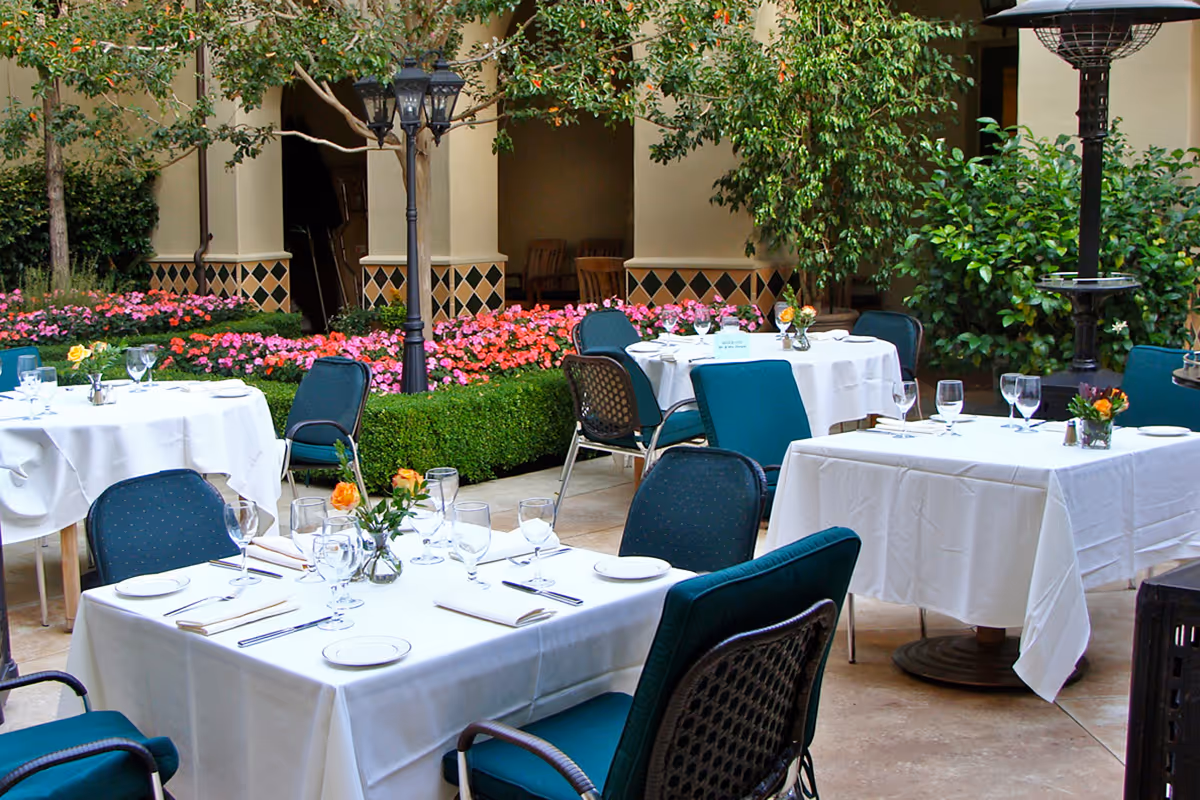 Outdoor dining area with tables covered in white tablecloths, set with glassware, plates, and silverware. The area is surrounded by green plants, colorful flowers, and decorative lampposts, creating a serene garden atmosphere.