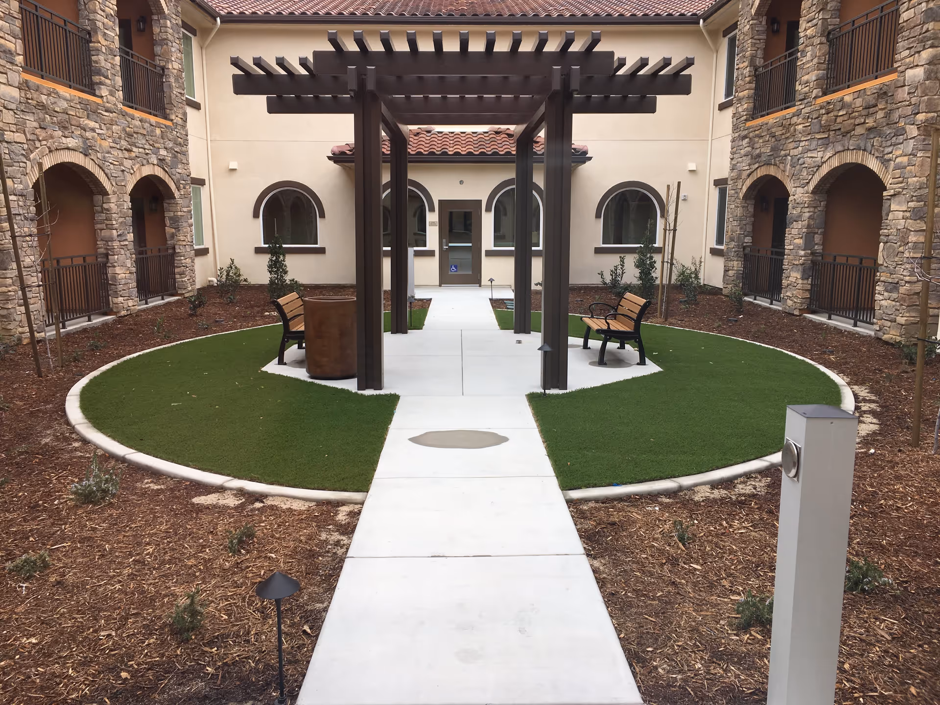 Outdoor courtyard area at Vineyard Ranch at Temecula featuring a concrete pathway leading to a pergola with benches on either side, surrounded by artificial grass and landscaped mulch beds. The courtyard is enclosed by a two-story building with stone and stucco walls and arched windows and doorways.