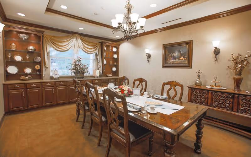 A traditional dining room with a long wooden table set for a meal, surrounded by eight wooden chairs. The room features built-in wooden cabinets displaying decorative plates and glassware, a window with draped curtains, a chandelier hanging from the ceiling, wall sconces, and framed artwork on the wall. There are also floral arrangements and decorative items on the table and sideboard.