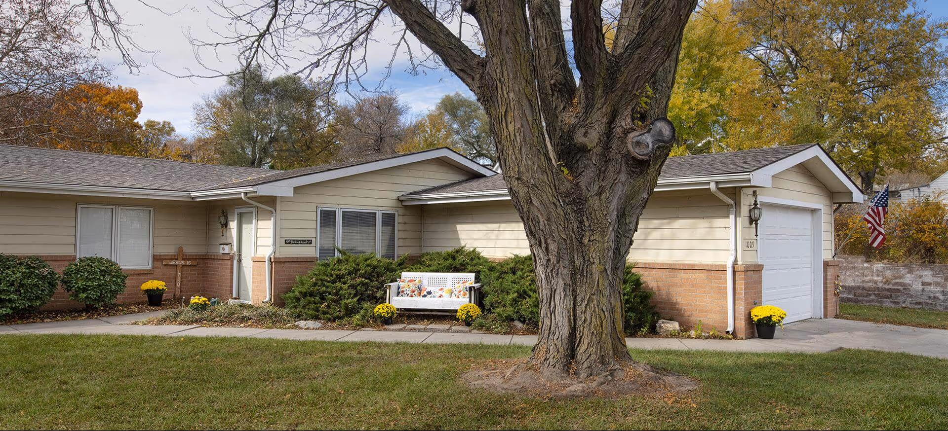 Single-story ranch-style building facade with a large tree in front, a white bench and potted flowers near the entrance and an American flag by the garage.