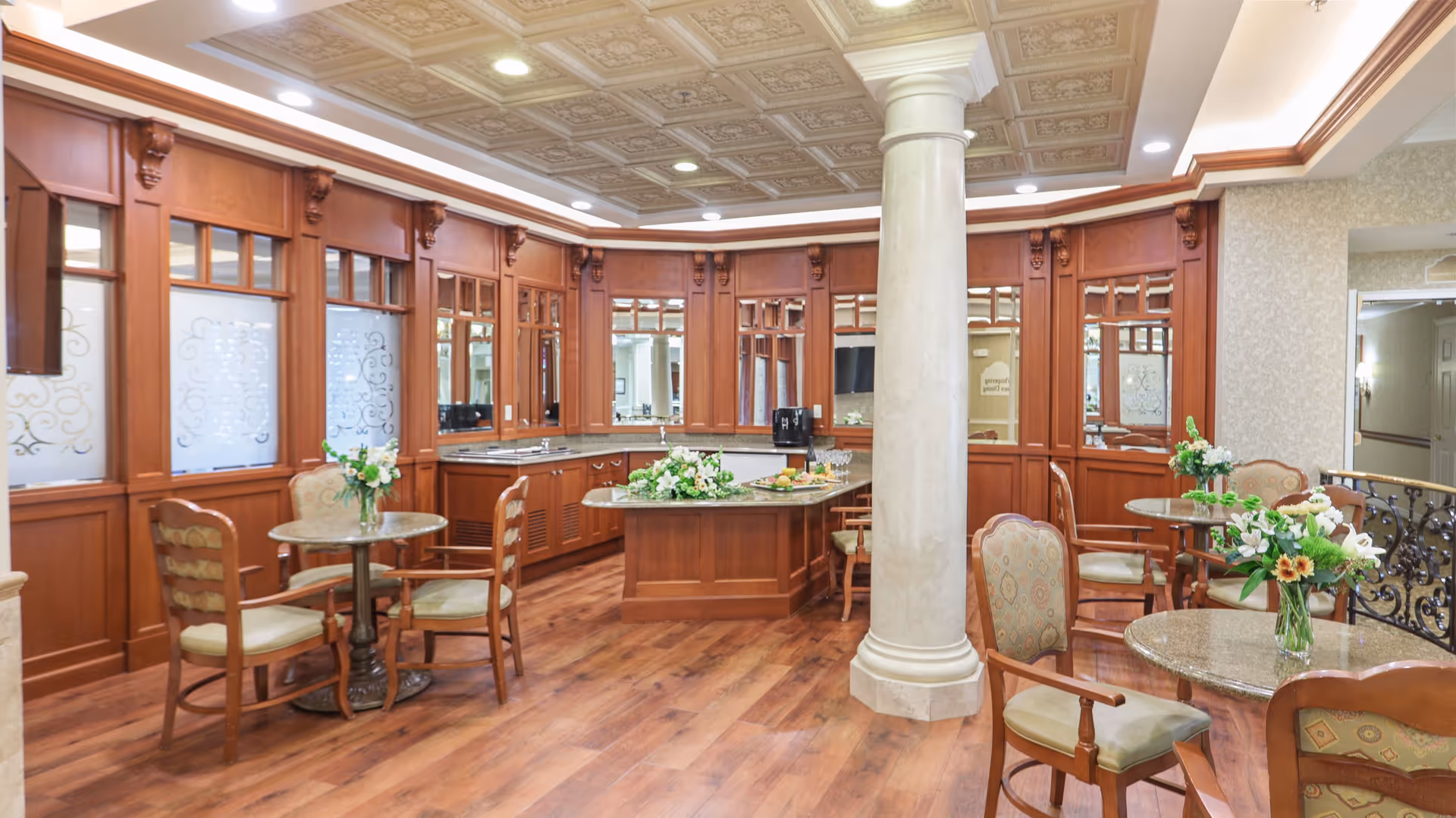 A spacious and elegant common area with wooden flooring, round tables with chairs, floral centerpieces, and a large central column. The room features wood-paneled walls with frosted glass windows and a decorative ceiling with recessed lighting.