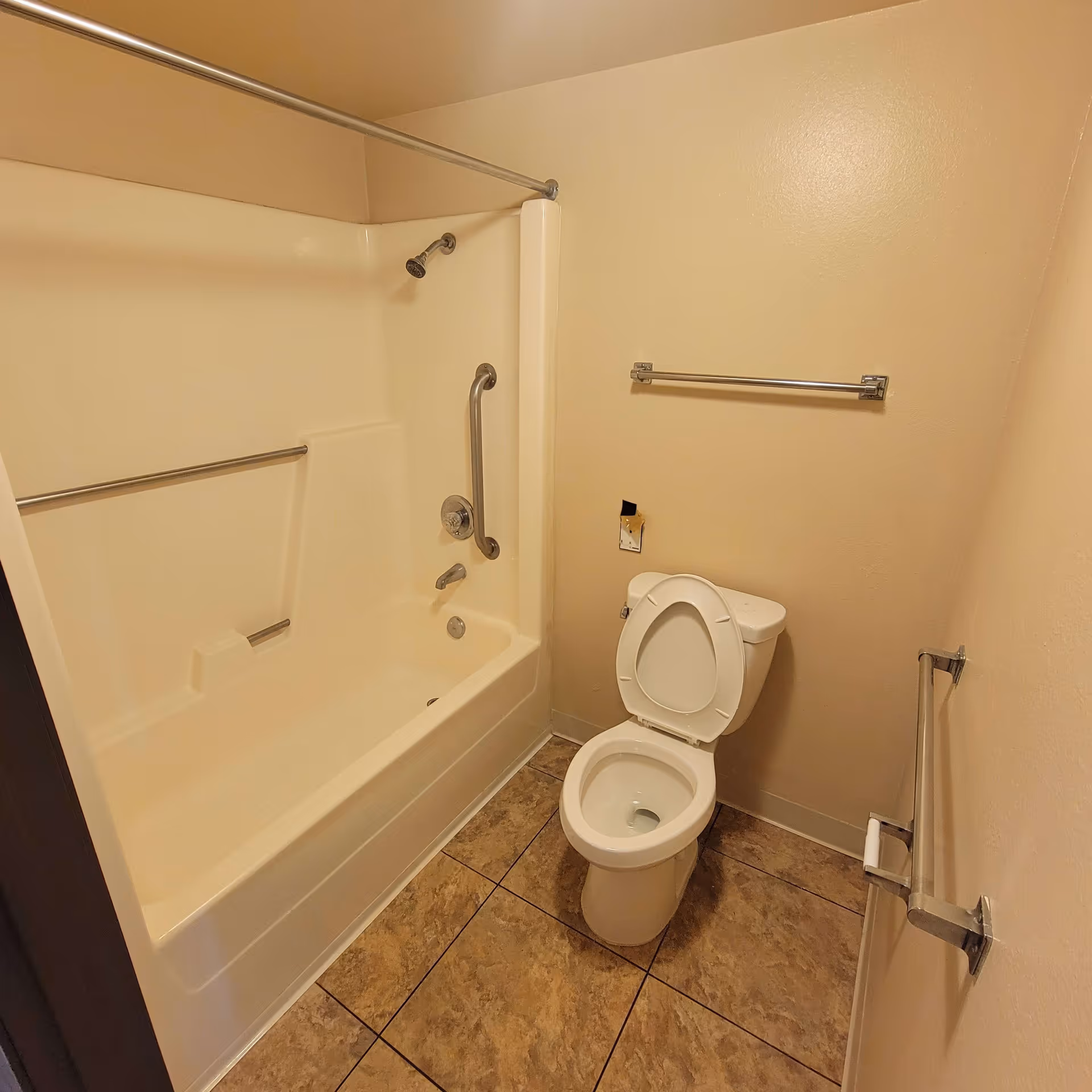 A small bathroom with beige walls and tiled floor featuring a white bathtub with a showerhead and grab bars, and a white toilet with the seat lid open. There are two towel bars on the walls, one above the toilet and one on the adjacent wall.