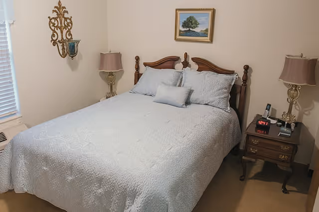 A neatly made bed with a white quilt and pillows in a bedroom. There are two bedside tables with matching lamps on each side of the bed. A small framed painting of a tree hangs on the wall above the bed. The room has beige walls and carpet, with a window covered by white blinds on the left side.