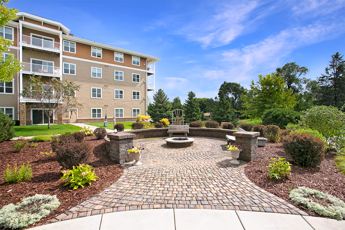 Front exterior of a multi-story senior living building with a landscaped circular brick courtyard and bench fountain.