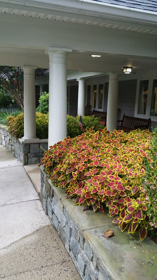 Covered outdoor walkway with white columns and stone planters filled with vibrant red and green foliage. Wooden benches are visible along the building wall under the covered area.