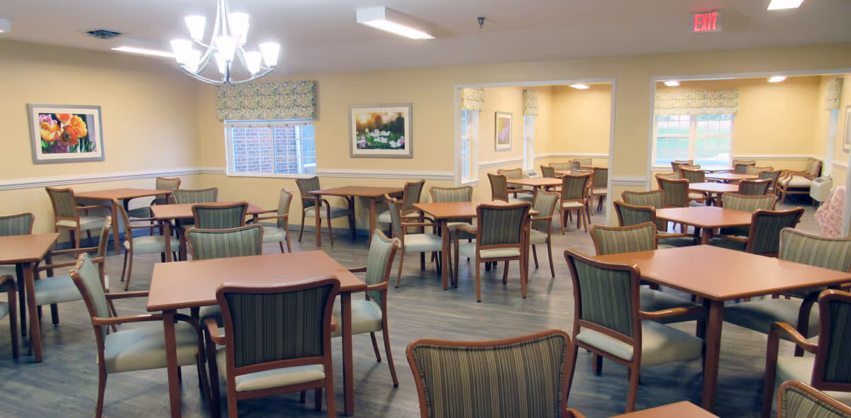 A spacious dining room with multiple wooden tables and chairs arranged neatly. The room has light yellow walls, framed floral artwork, windows with patterned valances, and a chandelier hanging from the ceiling. The floor is a dark wood finish, and there is an exit sign visible above a door in the background.