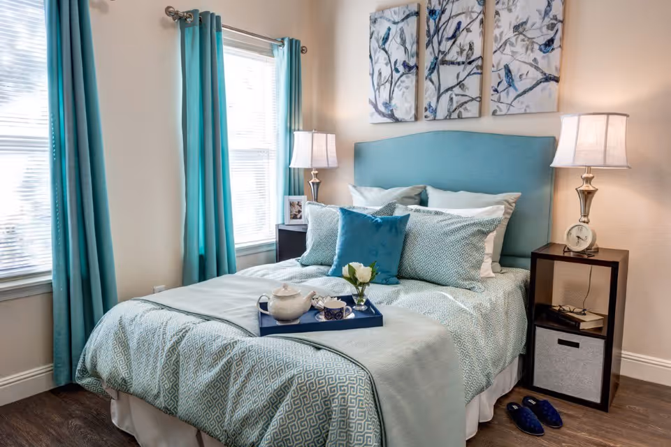 A neatly made bed with a teal upholstered headboard and matching teal and white bedding. There are multiple pillows arranged on the bed, a tray with a teapot, cup, and a small flower vase. Two windows with teal curtains are on the left side, and two bedside tables with lamps, a clock, books, and glasses are on either side of the bed. Blue slippers are placed on the floor next to the right bedside table. Three framed artworks featuring birds on branches hang above the headboard.