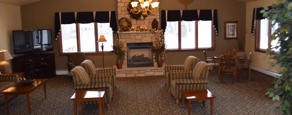 A cozy common room with a stone fireplace, striped armchairs, side tables, a TV, and a small dining table by the windows.