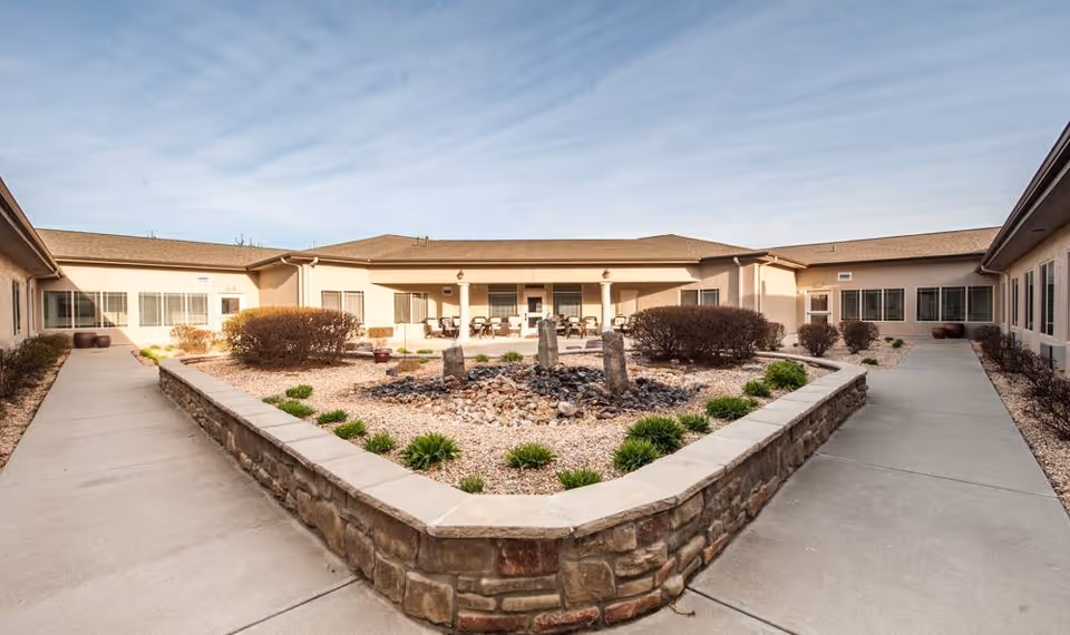 Sunlit central courtyard with a raised stone planter and rock fountain, surrounded by walkways and a single-story senior living building with patio seating.