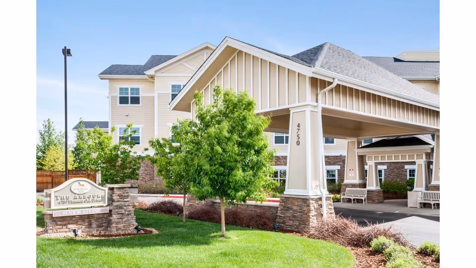 Exterior view of a senior living facility named The Arbours at 4750 Pleasant Oak Drive, featuring a covered entrance with stone pillars, green trees, and well-maintained landscaping under a clear blue sky.