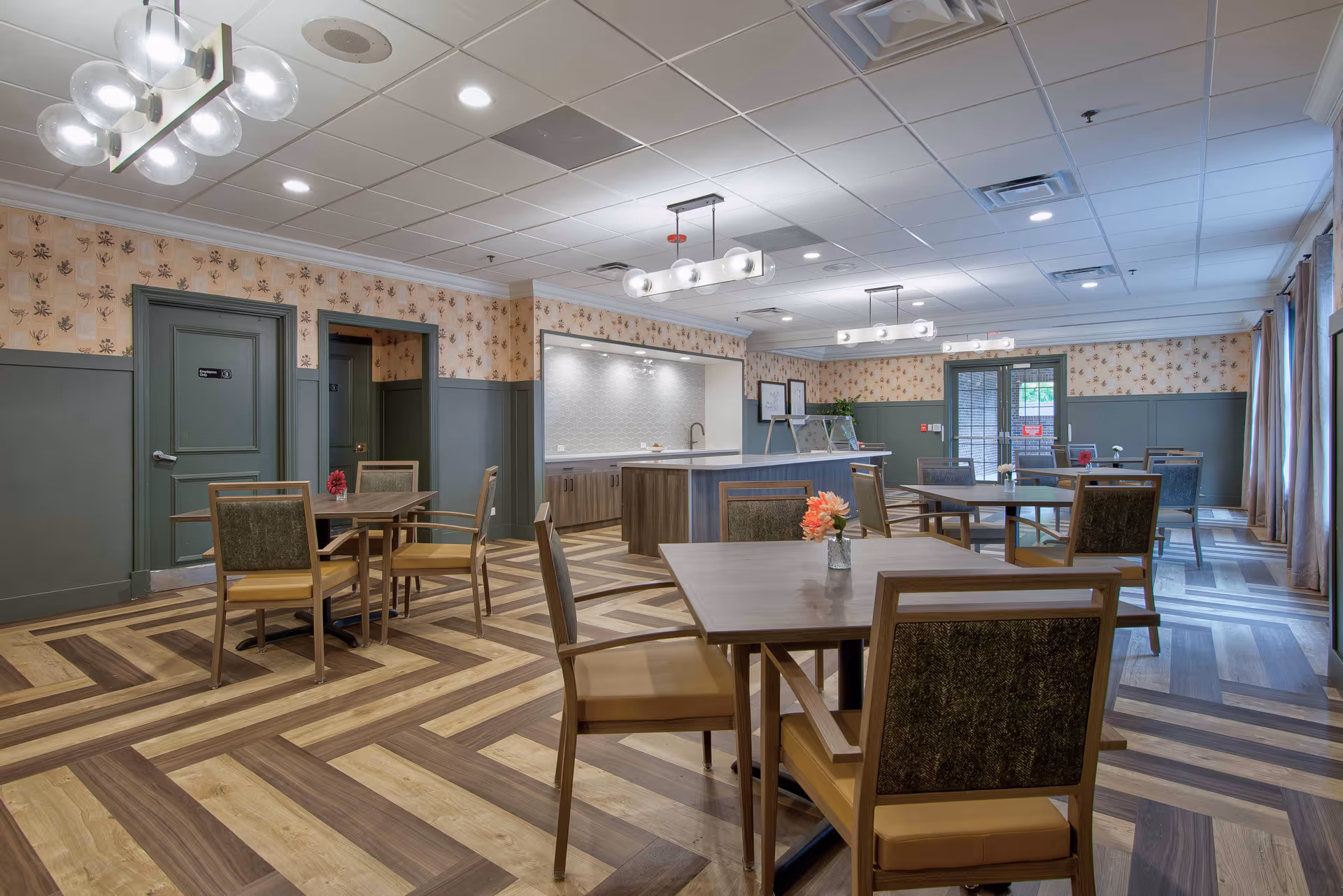 A well-lit dining room with several wooden tables and chairs arranged neatly. The room features patterned wallpaper with a floral design on the upper half of the walls and green paneling on the lower half. The floor has a geometric wood pattern. Modern light fixtures hang from the ceiling, and there are large windows with curtains on the right side. A counter area with cabinets and a sink is visible at the back of the room.