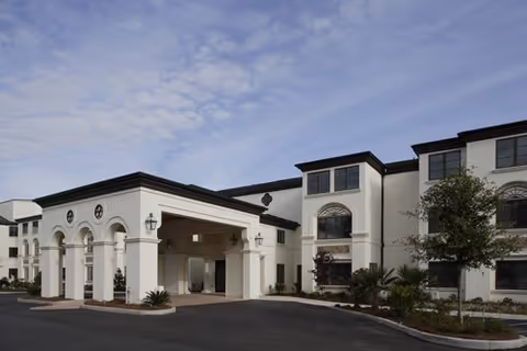 Exterior view of a large, white senior living facility building with multiple windows and a covered entrance driveway under a partly cloudy sky.