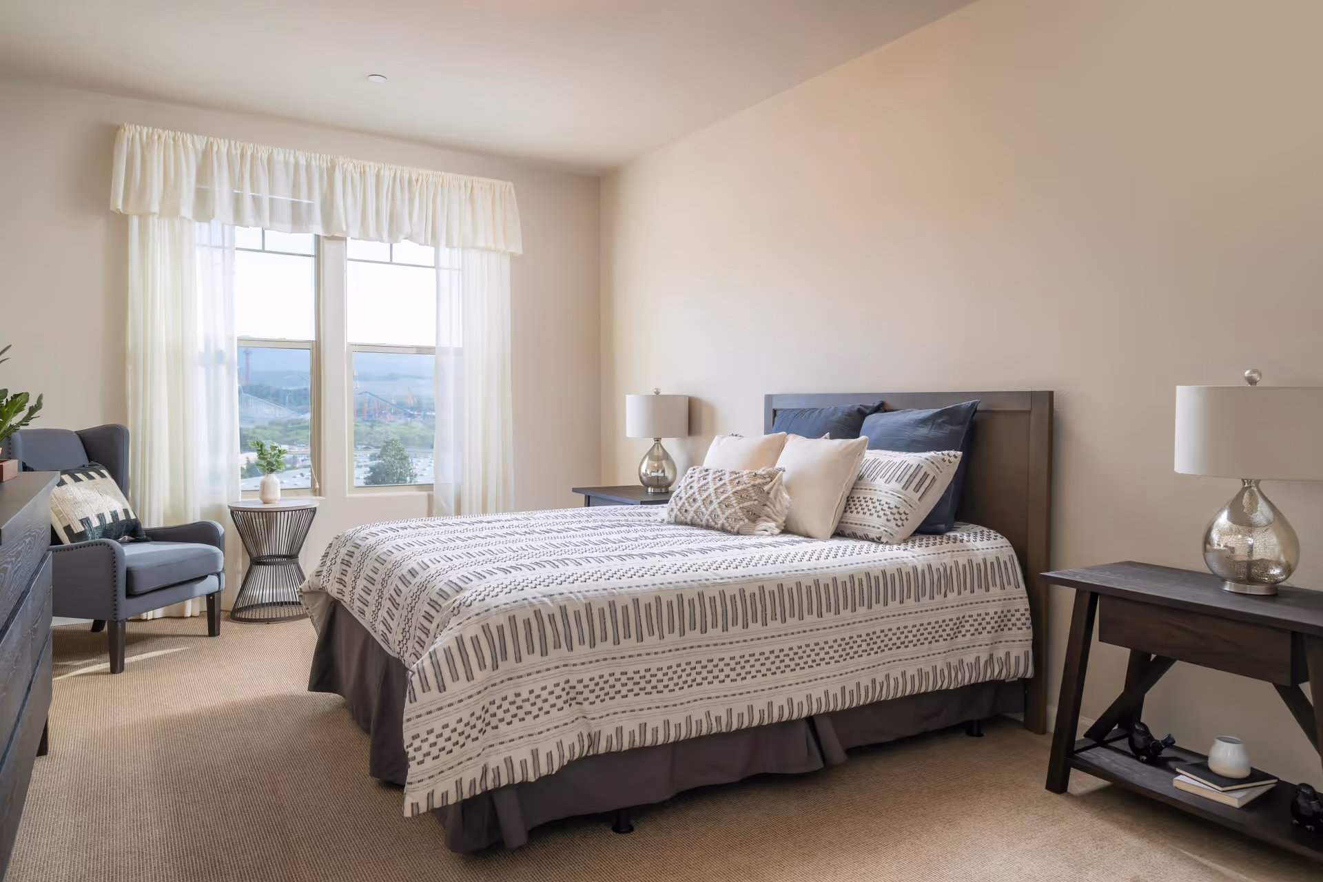 A bright bedroom with a large bed featuring patterned bedding and multiple pillows. There are two matching bedside tables with lamps on either side of the bed. A comfortable armchair with a decorative pillow sits near a window with sheer white curtains, allowing natural light to fill the room. The walls are painted a soft beige color and the floor is carpeted.