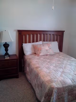 A small bedroom featuring a wooden headboard, a bed with a pastel patchwork quilt, and a nightstand with a lamp.