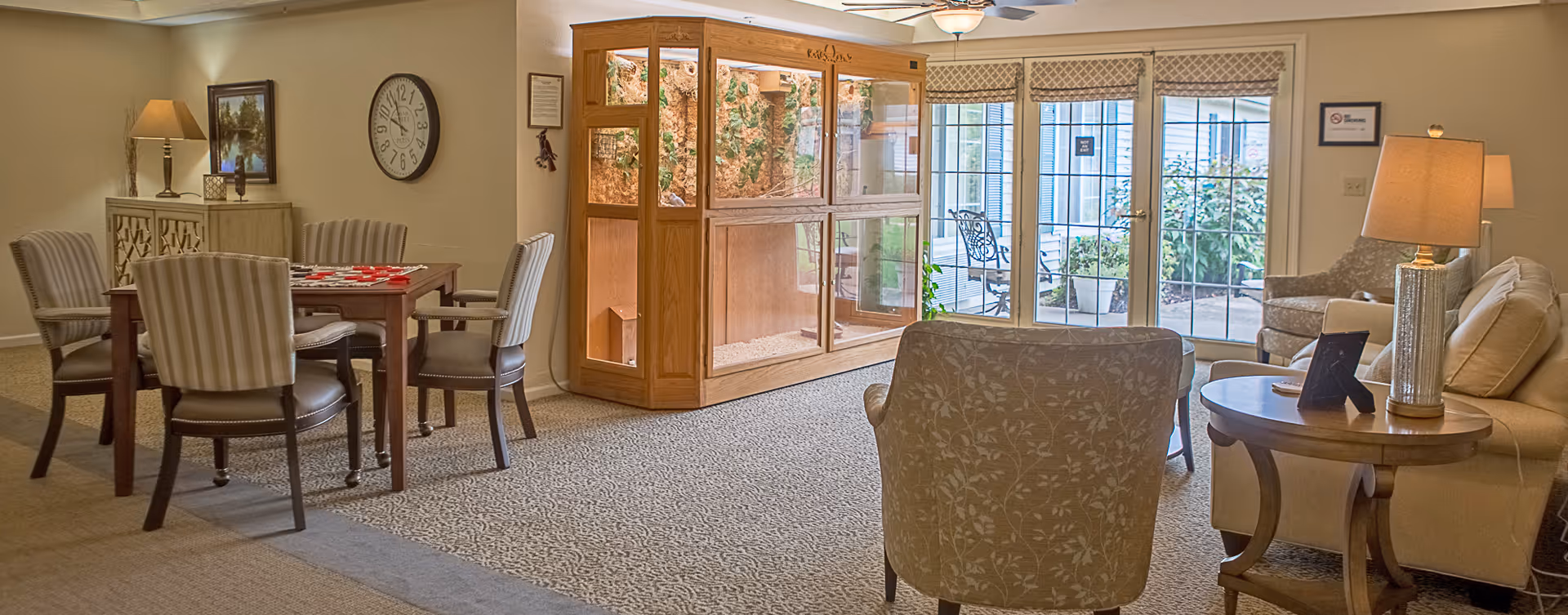 A cozy living area in a senior living facility featuring a small dining table with four striped chairs, a large wooden display cabinet with glass panels, a beige sofa, an armchair with floral upholstery, a round wooden side table with a lamp, and large windows overlooking an outdoor patio with plants.