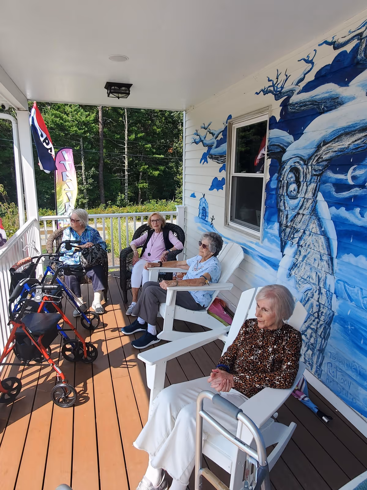 Four elderly women sitting on a covered wooden porch with white chairs and a mural of a tree and moon on the wall behind them. Two walkers are visible near the women, and there are trees and greenery in the background outside the porch.