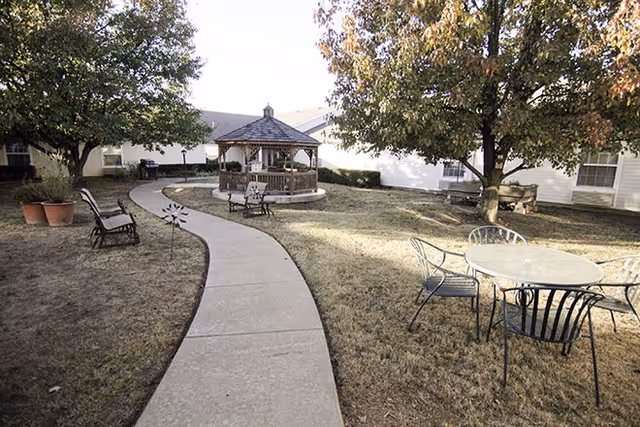 Outdoor garden area with a curved concrete walkway leading to a wooden gazebo. There are several metal chairs and tables scattered around the grassy area, along with a few trees providing shade.